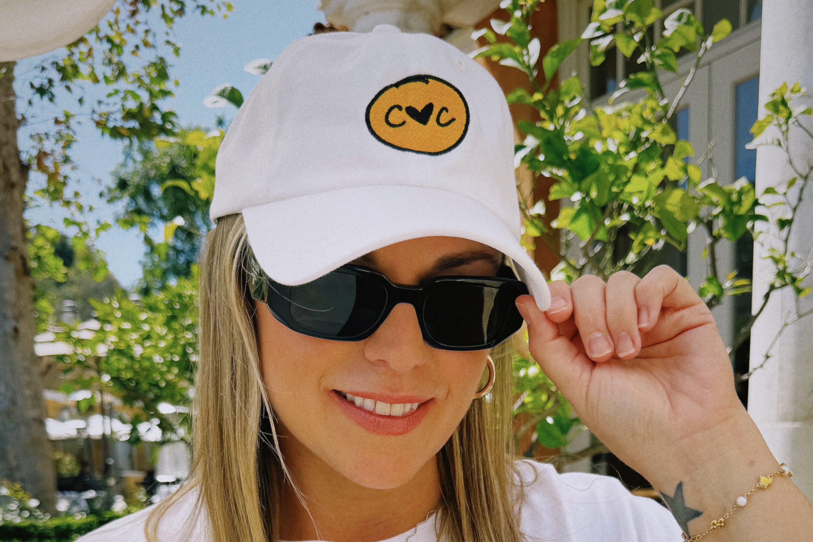 Woman wearing a white cap with a logo, sunglasses, and smiling outdoors.